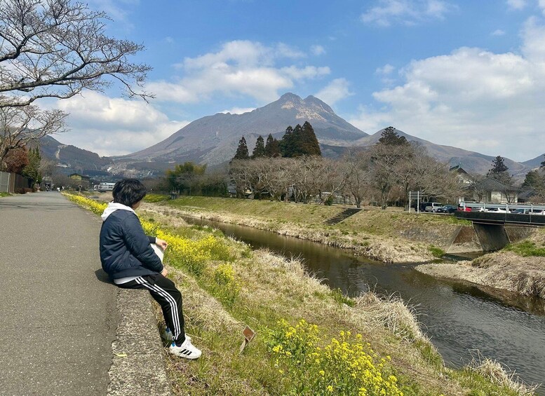 A person sitting by a river with mountains in the background. 1