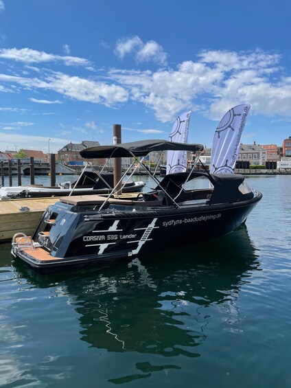 A black boat named 'CORSA 595 tender' docked at a marina with a cityscape in the background. 3