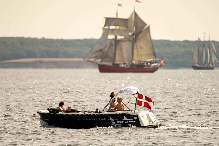 A small motorboat with two people and a flag, moving on water with two sailboats in the background. 1