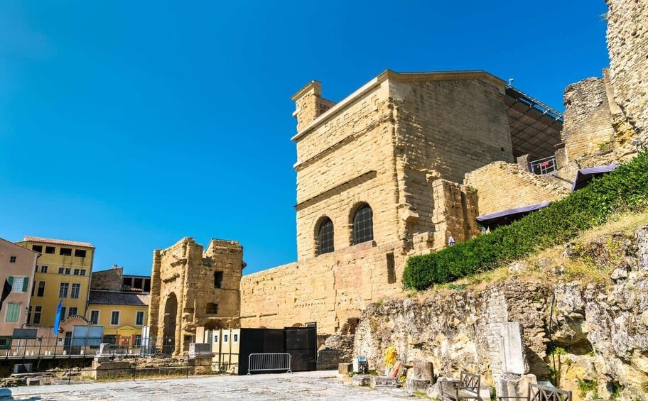 A historic stone building with arched windows and a tower, surrounded by modern buildings and a rocky hillside. 5