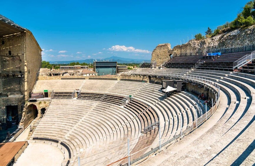 An ancient amphitheater with stone seating and a stage area, surrounded by modern buildings and a clear sky. 3