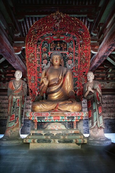 A large seated Buddha statue with intricate red and gold decorations, flanked by two smaller statues in traditional attire, set against a backdrop of a richly decorated temple interior. 2