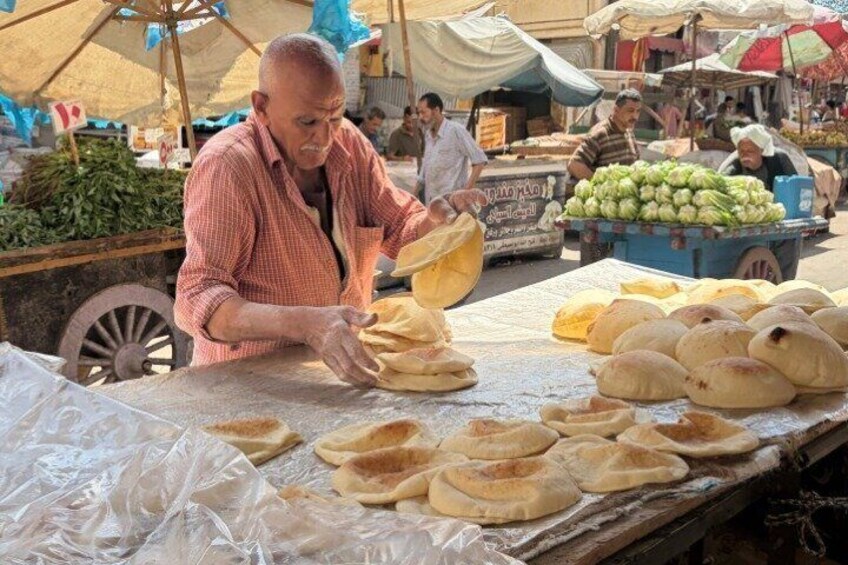 A street vendor preparing food at a market stall with various items and people in the background. 1