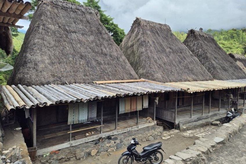 Thatched-roof huts with corrugated metal roofs, a motorcycle, and a stone pathway. 2