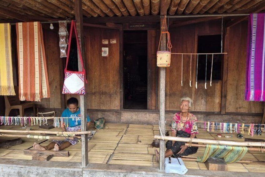 Two individuals engaged in weaving on a wooden porch with colorful textiles and tools. 4