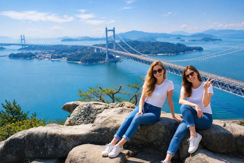 Two people sitting on rocks with a suspension bridge and a body of water in the background. 1