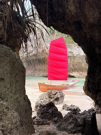 A wooden boat on a sandy beach with a red sail, viewed through a rocky cave entrance. 3
