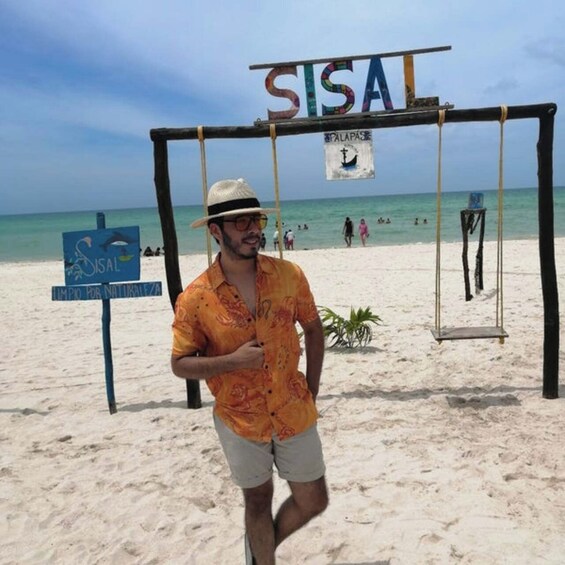 A man on a beach with a sign that reads 'SISAL' and another sign that says 'PALAPASE.' 1