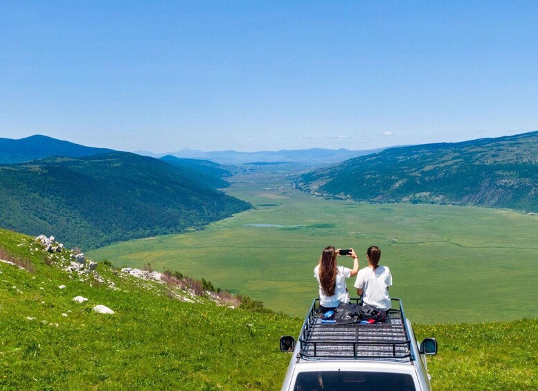 Two people sitting in a vehicle on a grassy hill overlooking a valley with mountains in the distance. 2