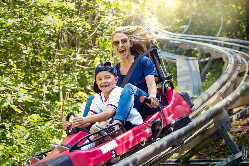 Two people on a red go-kart on a winding track surrounded by greenery. 1