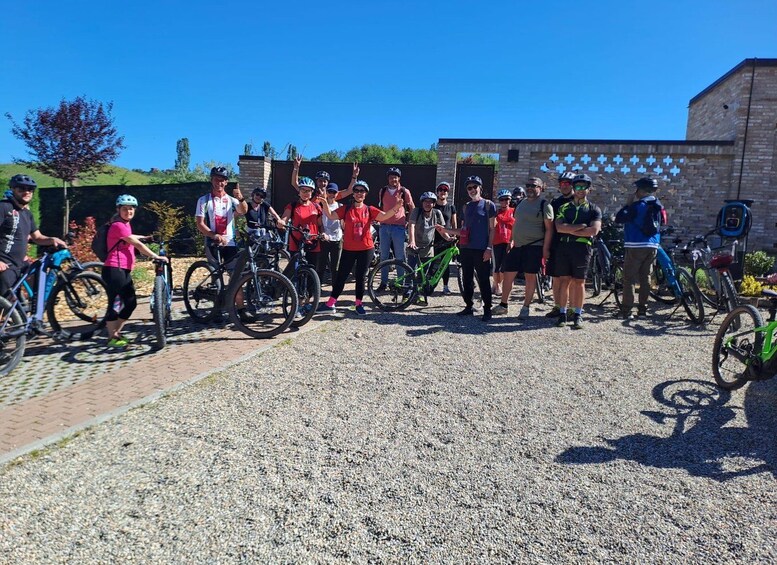 A group of cyclists gathered outdoors, some with bicycles, in front of a building with a distinctive wall pattern. 4