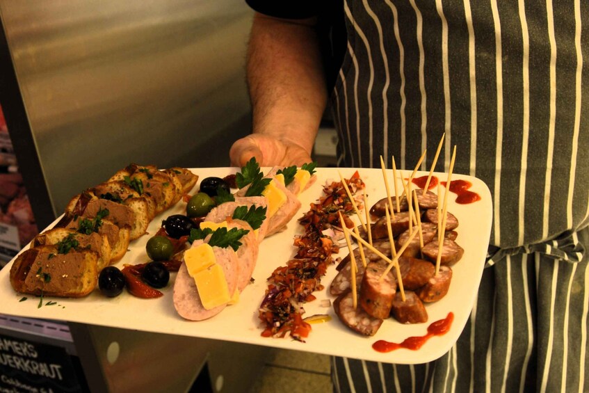A person holding a tray with various food items including bread, olives, cheese, and skewered sausages. 3