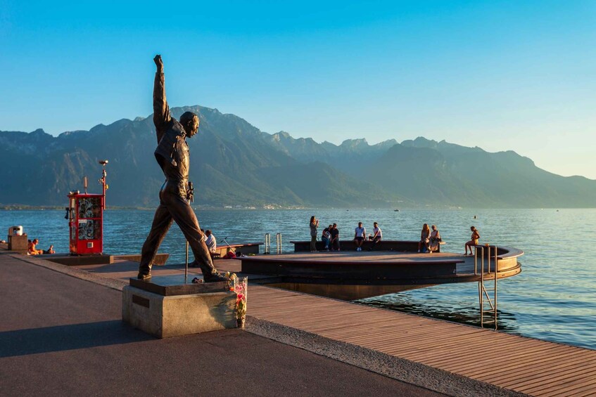A statue of a man with raised arms on a pier, a red kiosk, and mountains in the background. 1