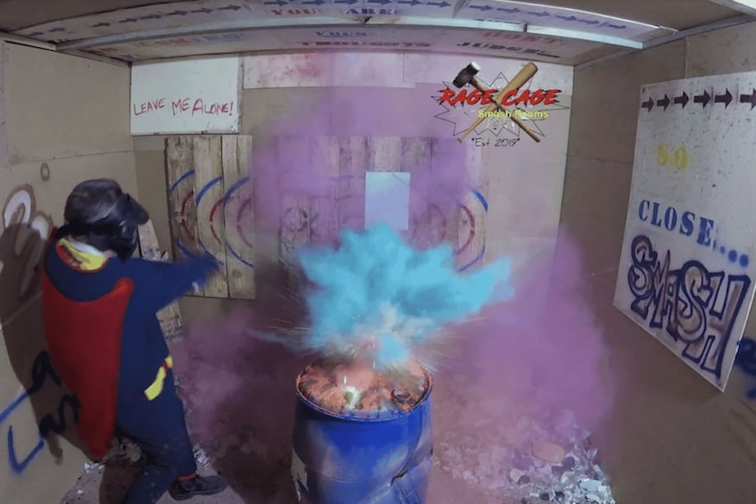 A person in a blue and red costume throwing a blue smoke bomb in a room with graffiti and a sign that says 'RAGE CAGE Smash Rooms.' 2