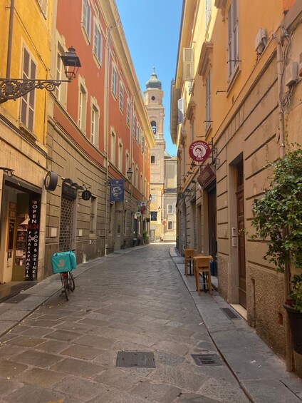 A narrow street with buildings on either side, a bicycle, and a clock tower in the distance. 3