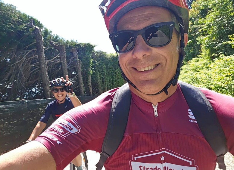 A person wearing a red cycling jersey and a helmet, taking a selfie with another cyclist in the background. 2
