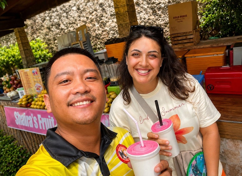 Two people holding pink drinks with straws, standing in front of a fruit stand with a sign that reads 'Shandra's Fruit.' 3