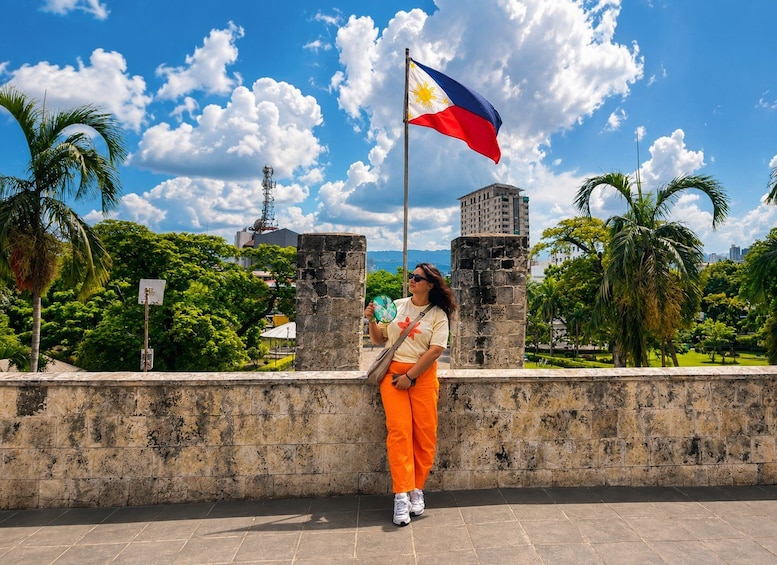 A person standing by a stone wall with a Philippine flag, palm trees, and a clear blue sky. 5