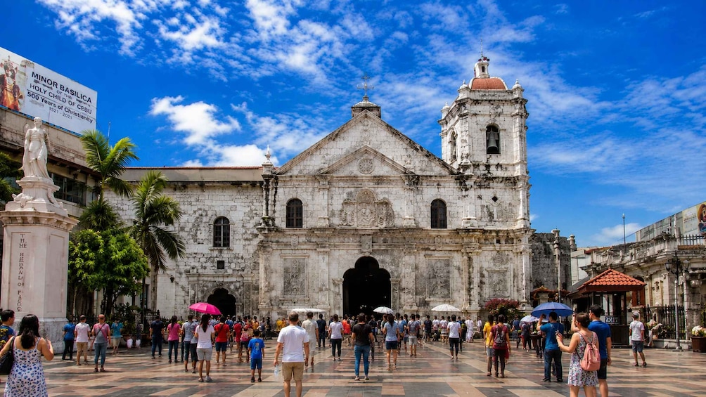 A historic church with a bell tower, surrounded by palm trees and people. 2