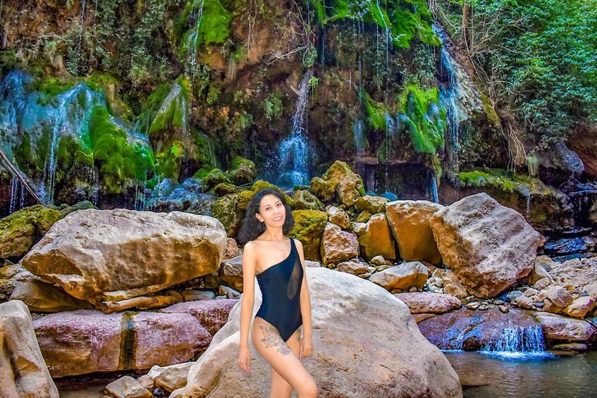 A person in a swimsuit standing in front of a waterfall with rocks and greenery. 3
