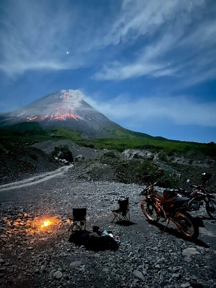 A volcanic landscape at dusk with a lit campfire, a motorcycle, and a chair. 1