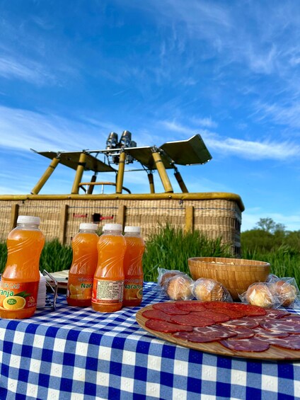 A picnic setup with a checkered tablecloth, bottles of orange juice, a wooden bowl, and sliced salami. 2