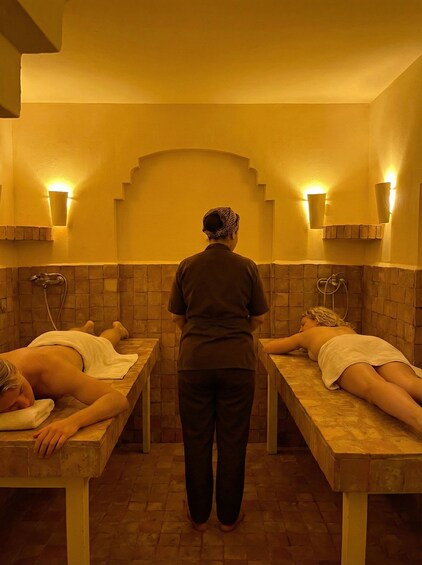 A spa room with two massage tables, a person standing between them, and wall-mounted lighting. 3