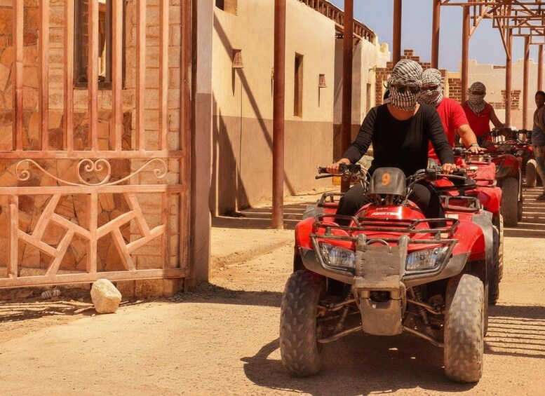 A person riding a red ATV on a dirt path, with buildings and a gate in the background. 2