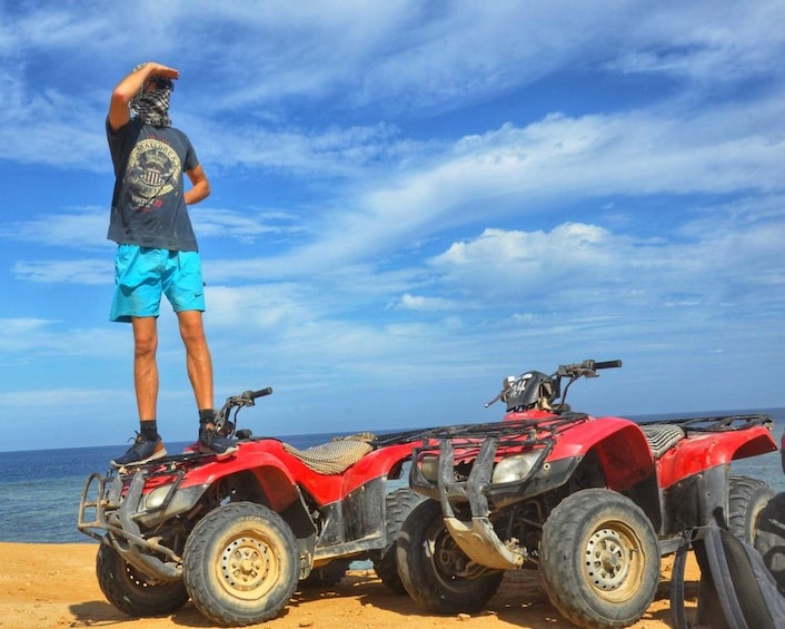 A person standing on a red ATV on a sandy beach with the ocean in the background. 3