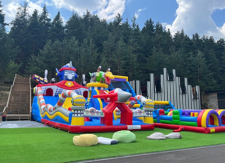 A colorful inflatable play structure with various slides and climbing areas, set against a backdrop of trees and a wooden fence. 1