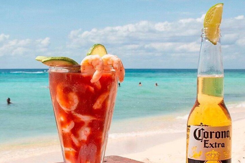 A beach scene with a glass of red drink with shrimp and lime, and a bottle of Corona Extra beer. 1