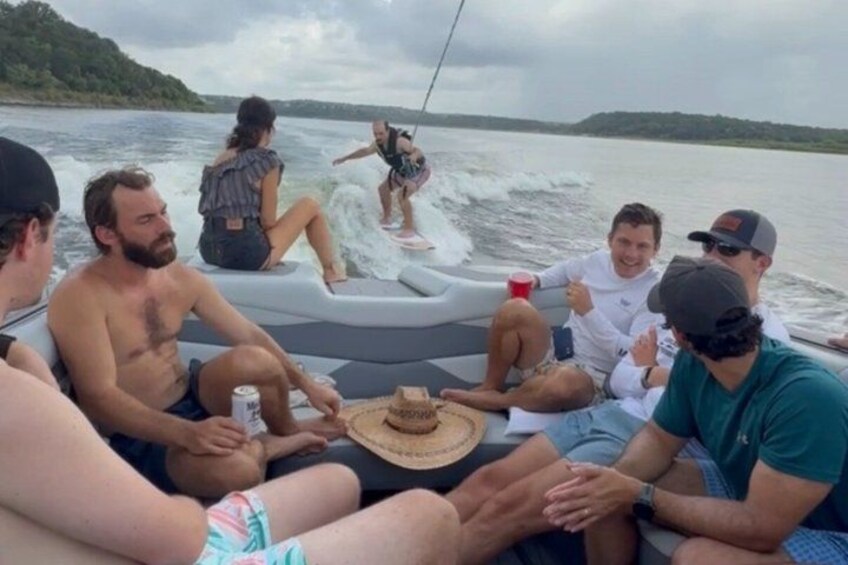 A group of people on a boat, some sitting and others standing, with a person surfing. 4