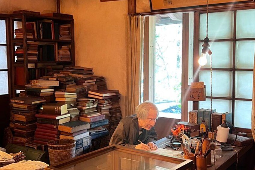 An elderly person writing at a desk with a large stack of books to the left and a window with curtains in the background. 4