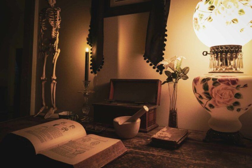 A dimly lit room with a skeleton, an open book, a bowl, a candle, and a floral lamp. 1