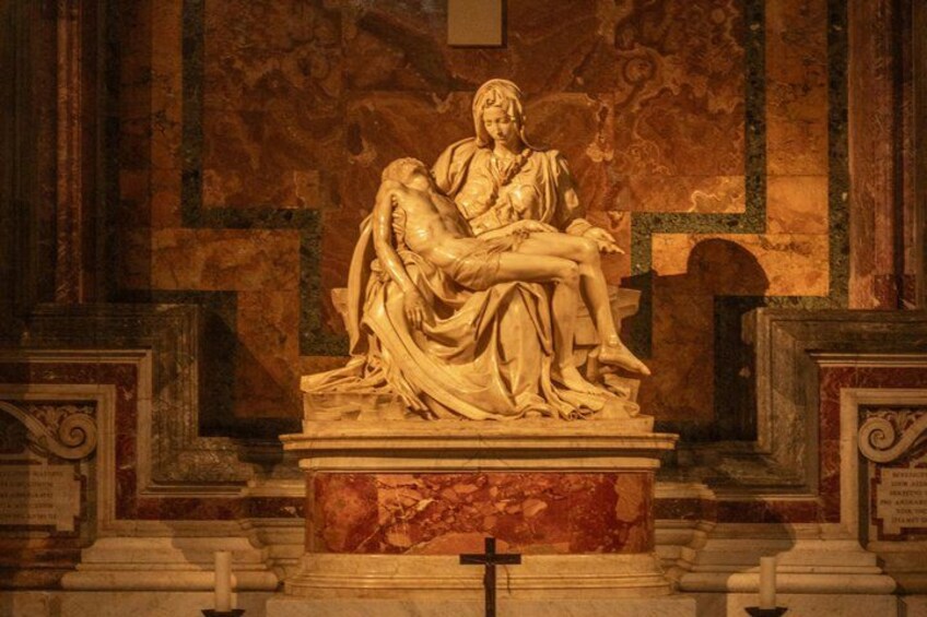A marble sculpture of a reclining figure with a child, set against a wall with a cross at the base. 4