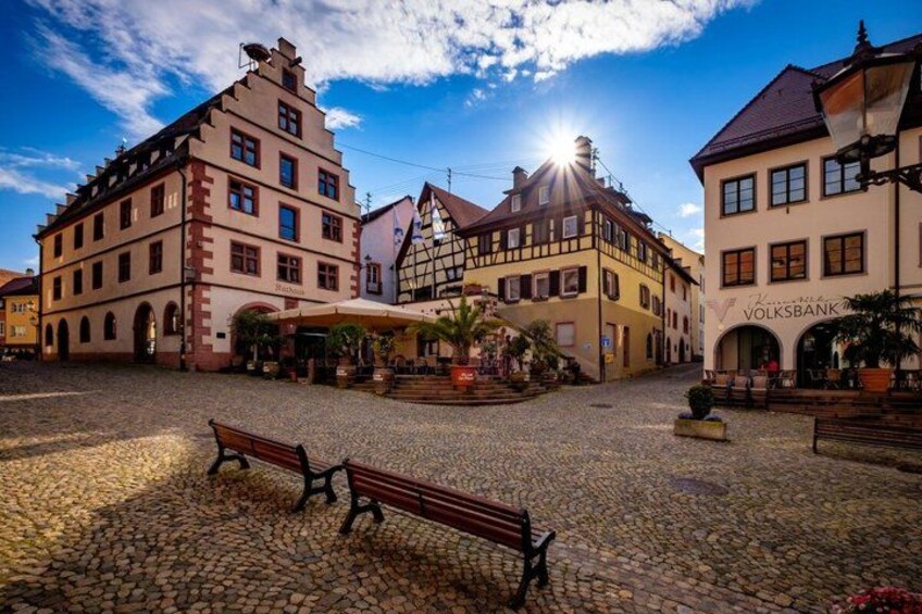 A cobblestone square with historic buildings, a bench, and a sunlit sky. 5