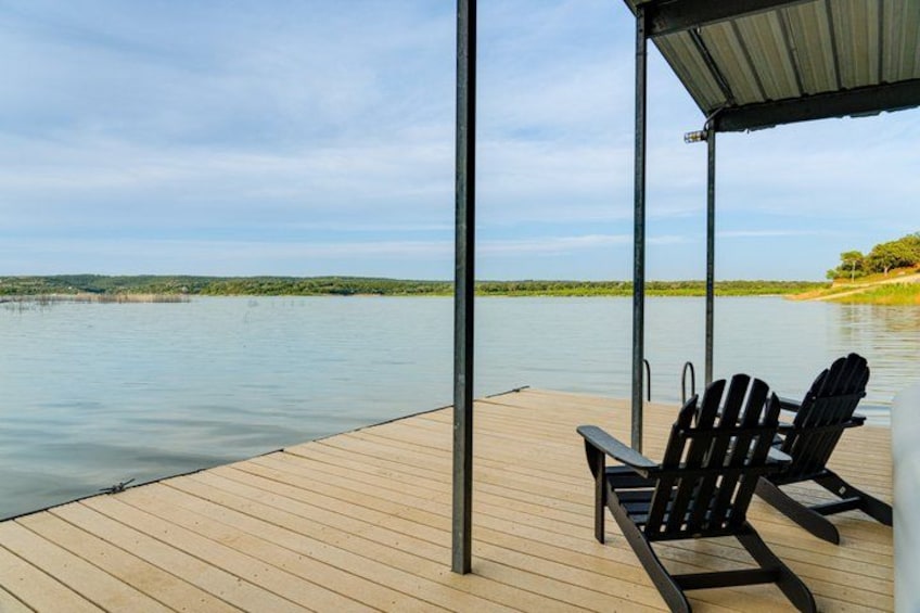 A wooden dock extending over calm water, with two black Adirondack chairs under a covered area. 4