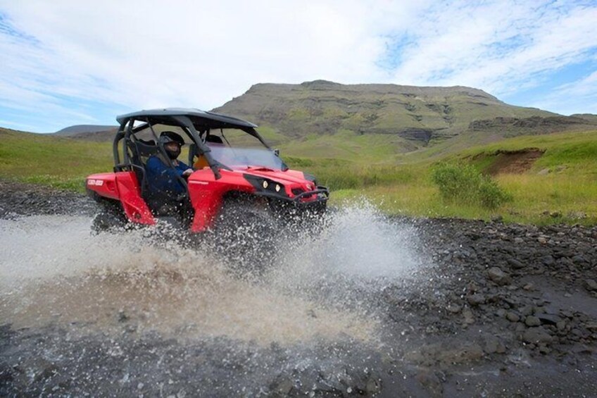 A red vehicle driving through a puddle, splashing water, with a hilly landscape in the background. 4