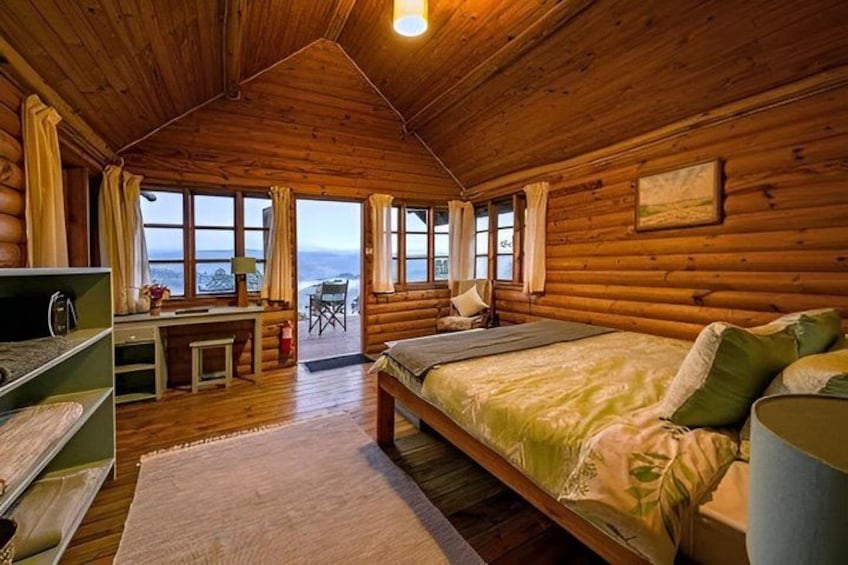 A wooden cabin interior with a bed, desk, and large windows. 4