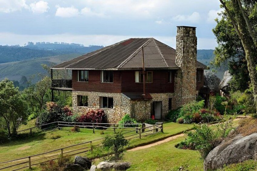 A two-story house with a stone chimney, surrounded by a lush garden and a scenic hillside view. 2