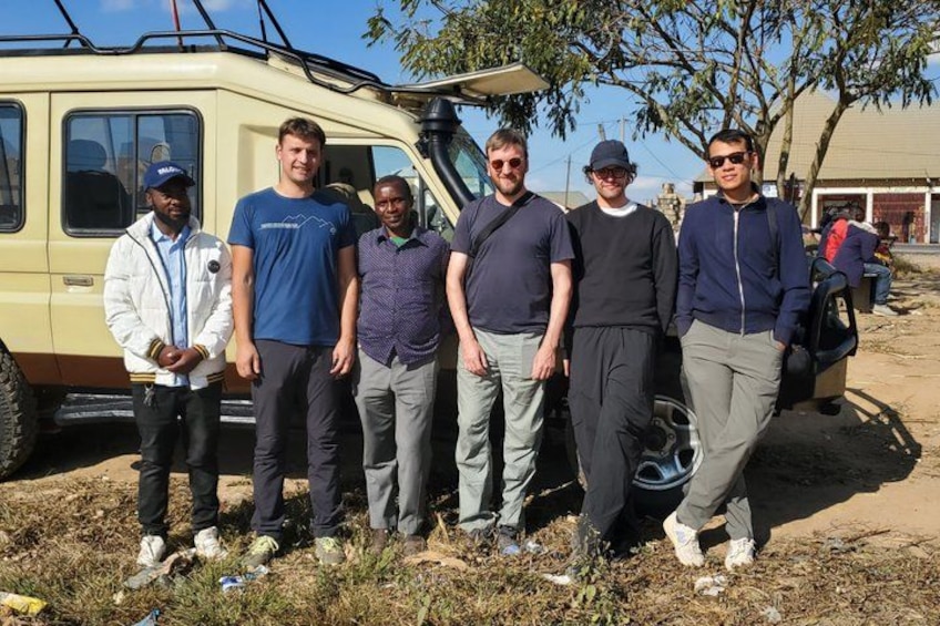 A group of people standing in front of a yellow vehicle, possibly a van, in a dry, grassy area with sparse trees. 1