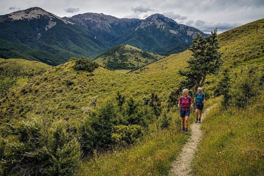 A hiking trail with two hikers, surrounded by green hills and mountains. 3