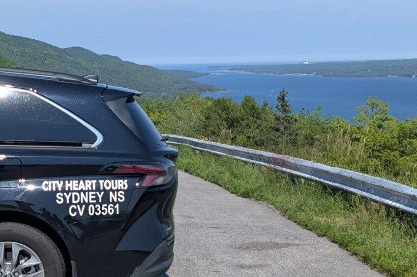 A black SUV with 'CITY HEART TOURS SYDNEY NS CV 03561' on the back, parked on a road with a guardrail and a scenic view of a body of water and hills. 1