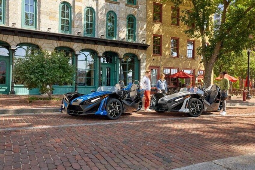 Two sports cars parked on a brick street in front of a building with green awnings and arched windows. 2