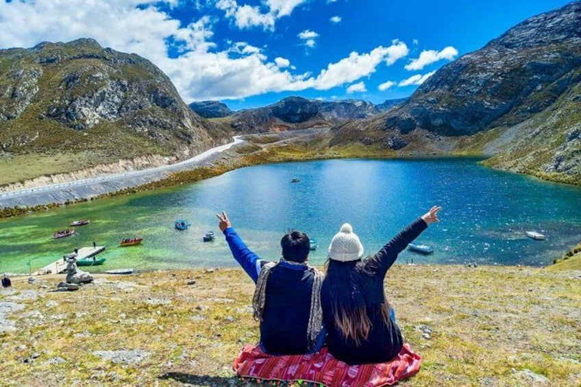 Two people sitting on a blanket overlooking a lake with boats, surrounded by mountains. 2