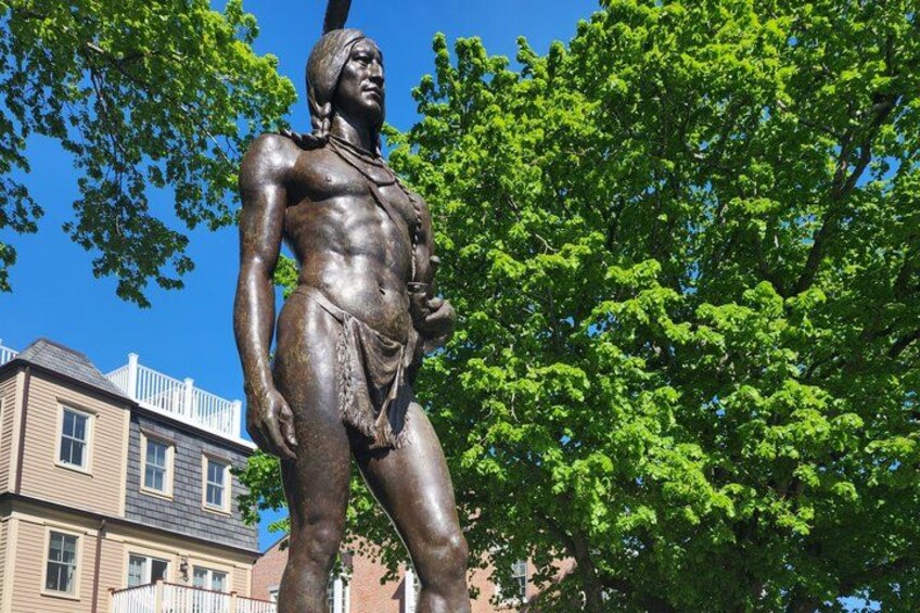 A bronze statue of a Native American figure with a feathered headdress, standing in front of a tree and a building. 2