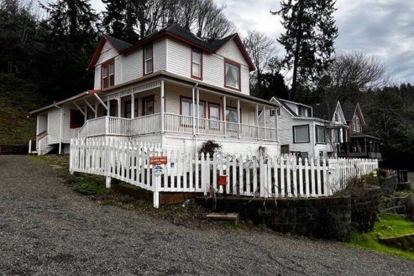 A two-story house with a porch, surrounded by a white picket fence, set in a wooded area. 4