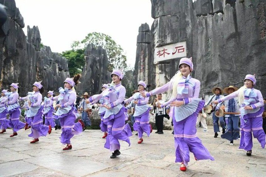 A group of dancers in purple costumes performing in front of a rocky landscape with a sign in the background. 3