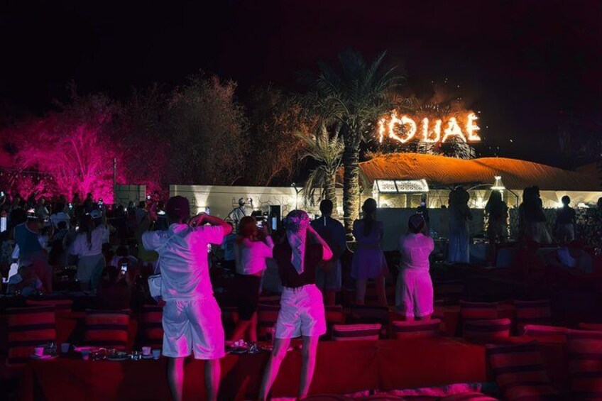 A nighttime outdoor event with a crowd, illuminated signage, and palm trees. 3