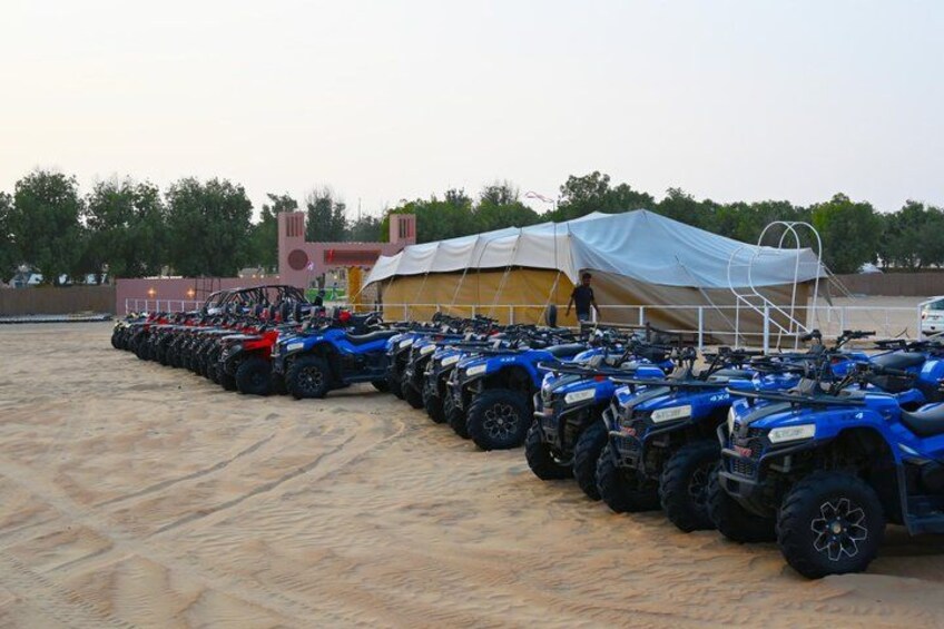 A row of ATVs parked in a sandy area with a tent and a building in the background. 2